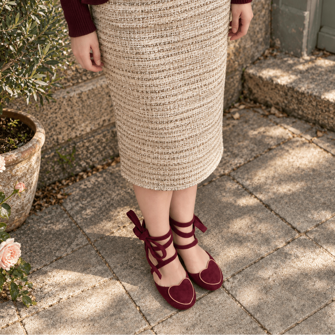 A person wearing a beige and brown textured knit pencil skirt with a dark burgundy top and matching burgundy heart-shaped lace-up shoes, standing on stone pavers next to a potted plant.