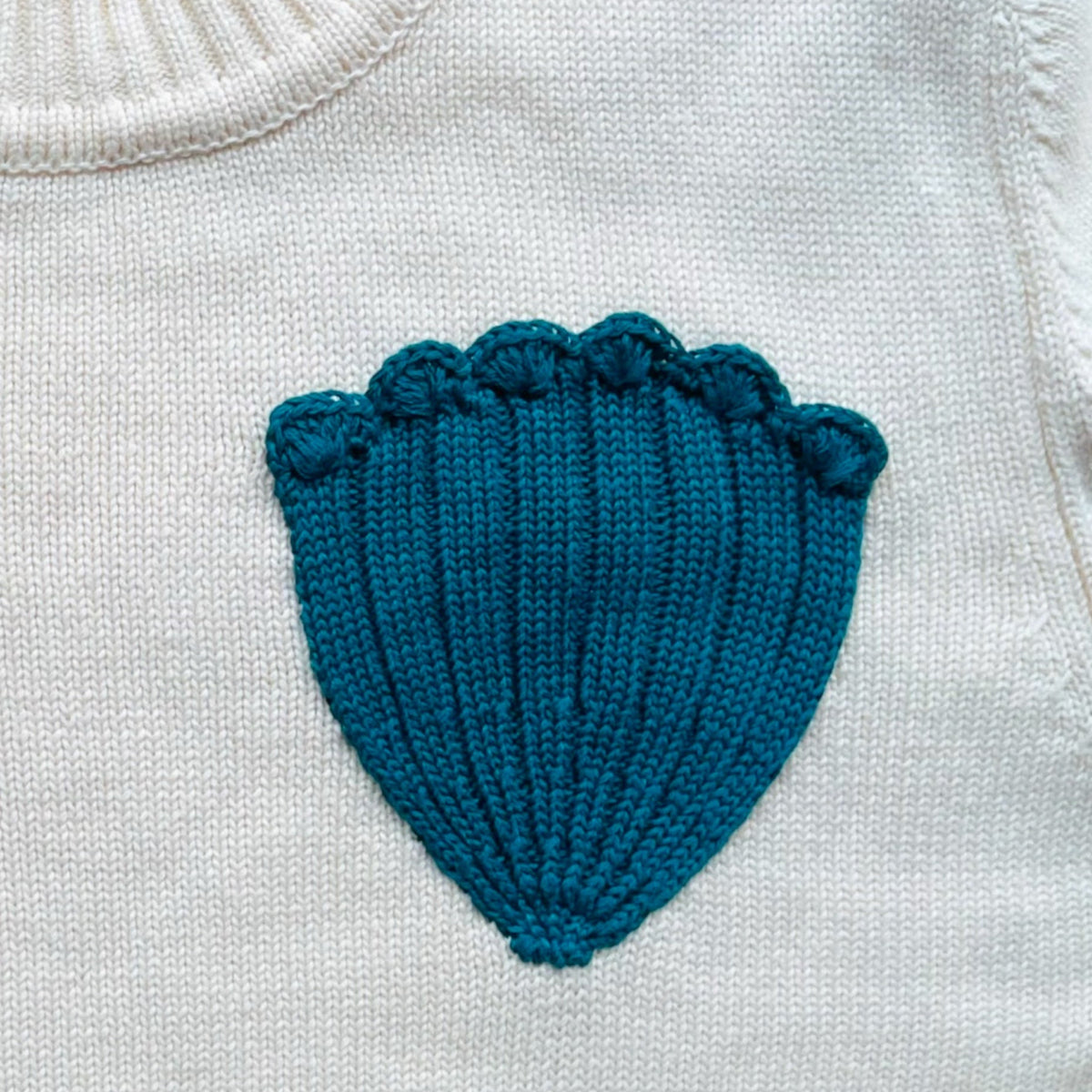 Close up of a turquoise knitted shell shape on an ivory jumper