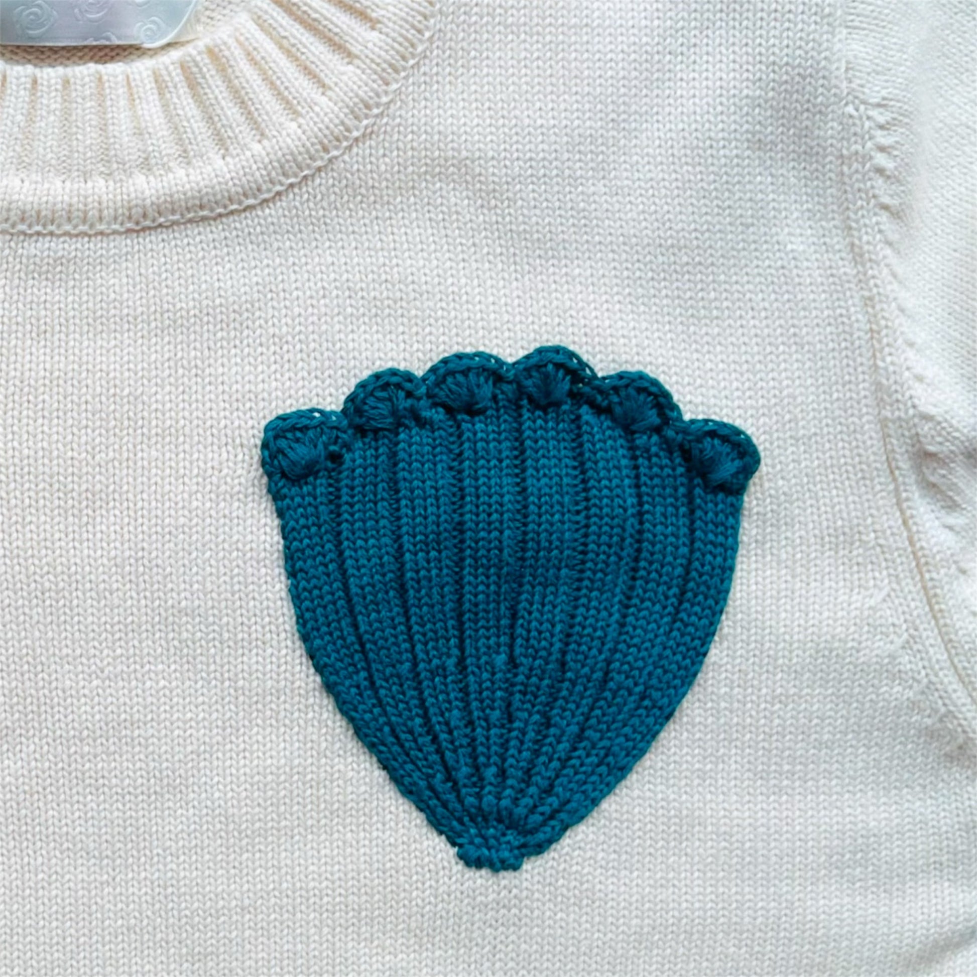 Close up of a turquoise knitted shell shape on an ivory jumper 