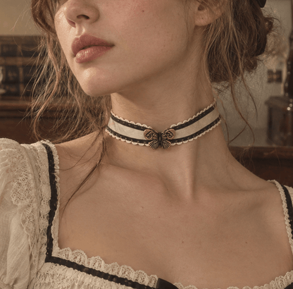 A woman in a vintage-style cream lace dress wearing a cream choker with black trim and a central bronze butterfly detail.