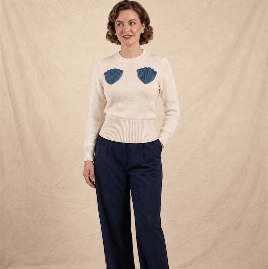 A woman stands against a beige fabric backdrop wearing a cream knit sweater with two blue shell-shaped crochet on the chest, navy wide-leg trousers.