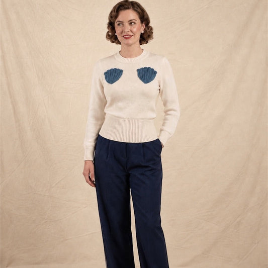 A woman stands against a beige fabric backdrop wearing a cream knit sweater with two blue shell-shaped crochet on the chest, navy wide-leg trousers.