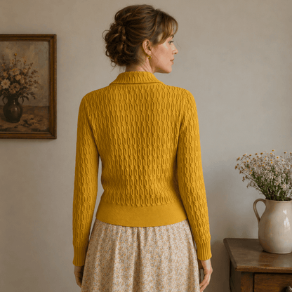 Woman wearing a belted mustard yellow cardigan and beige skirt standing in a room with a vase and painting on the wall.