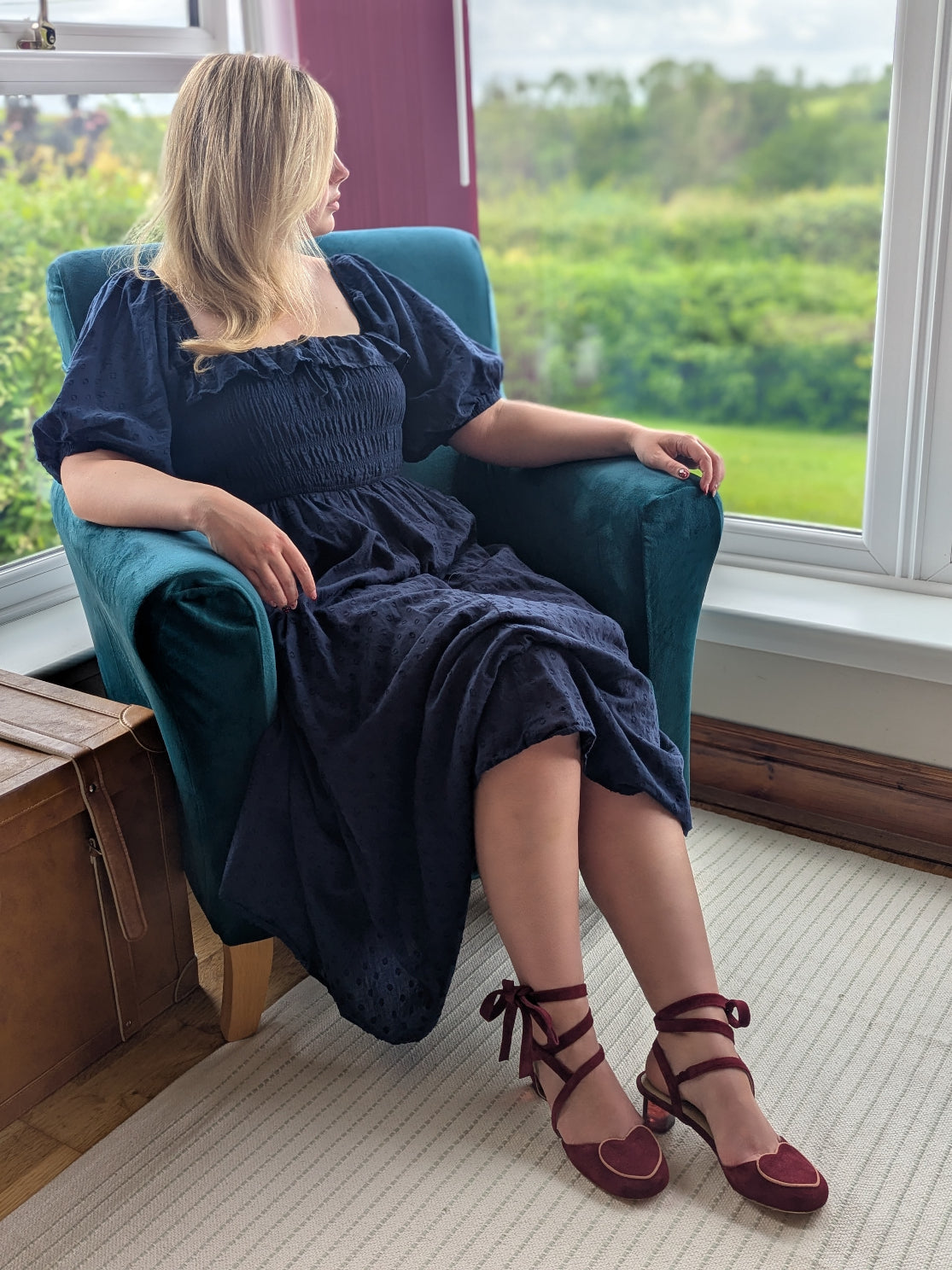 Woman in a dark blue dress sitting on a teal chair wearing burgundy ankle tie shoes