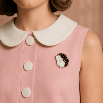 A close-up of a person wearing a pink sleeveless top with a white Peter Pan collar and three white buttons, featuring a small hedgehog brooch on the chest. The background is a soft pink wall with a blurred vase of flowers.