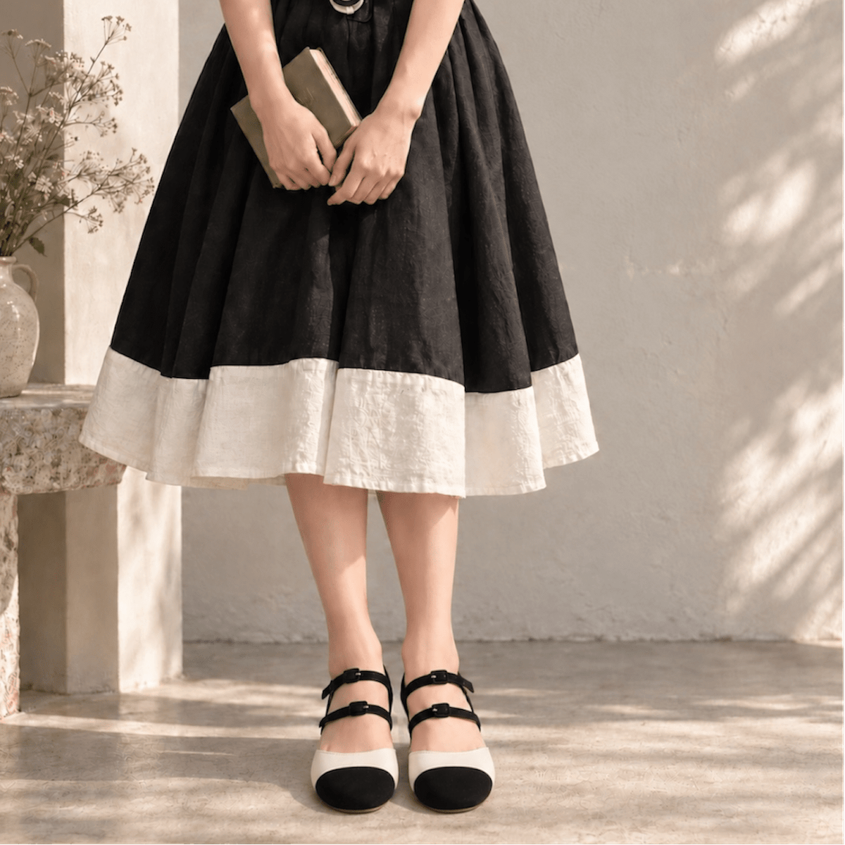 A person wearing a black and cream tiered linen skirt with black and cream Mary Jane shoes, holding a book in a sunlit, rustic interior setting.