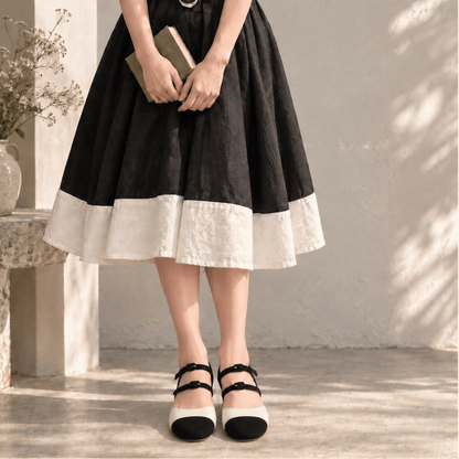 A person wearing a black and cream tiered linen skirt with black and cream Mary Jane shoes, holding a book in a sunlit, rustic interior setting.