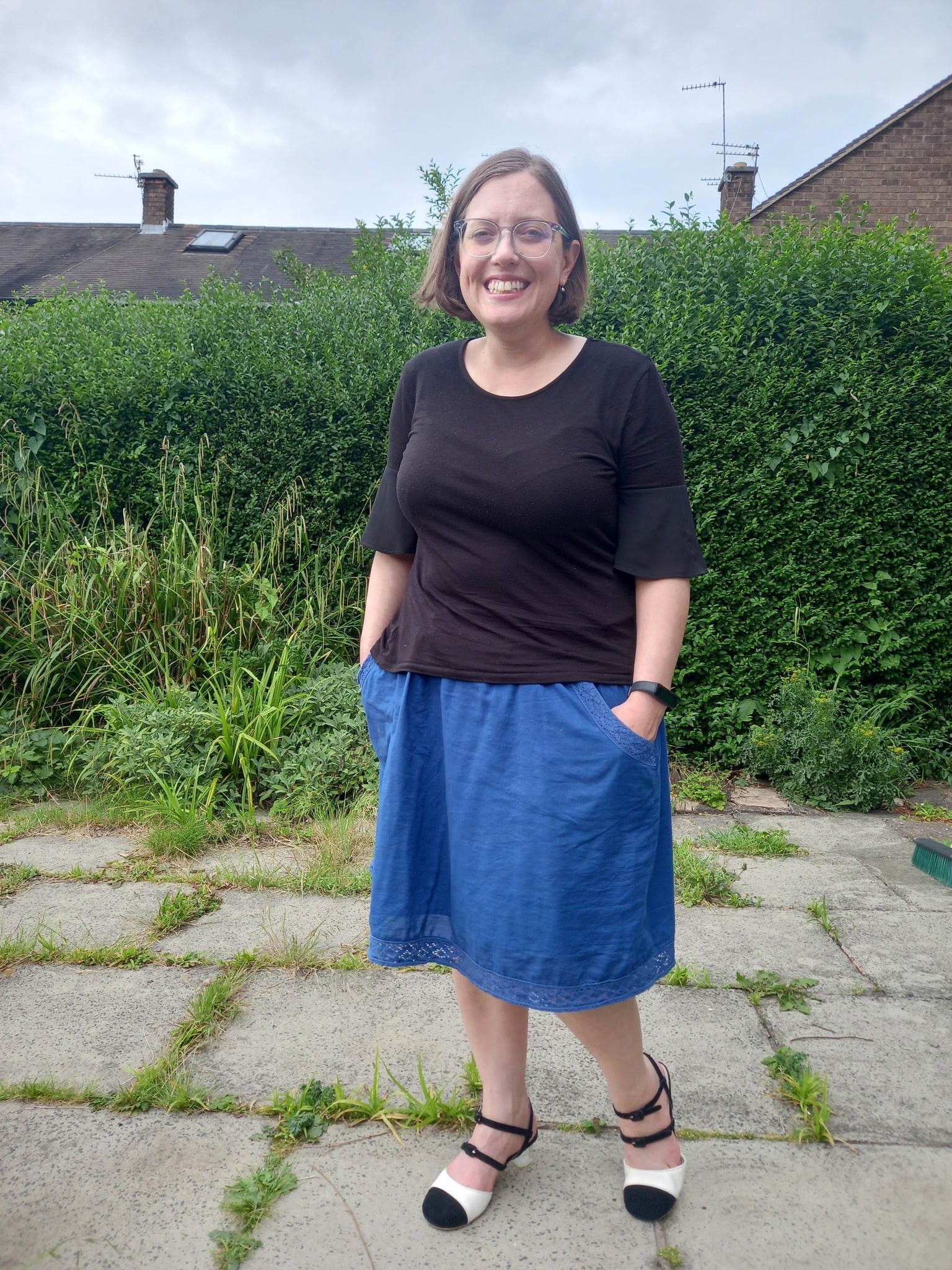 Woman wearing black and white Mary Jane shoes standing outdoors with greenery and a building in the background.